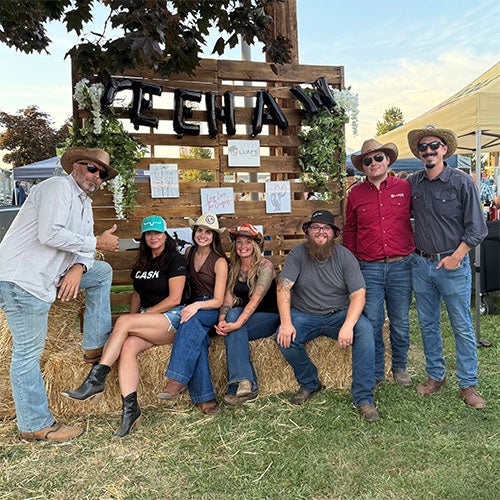 Six people in cowboy attire pose in front of a wooden pallet backdrop with black 'CHAH' balloons and decorations.
