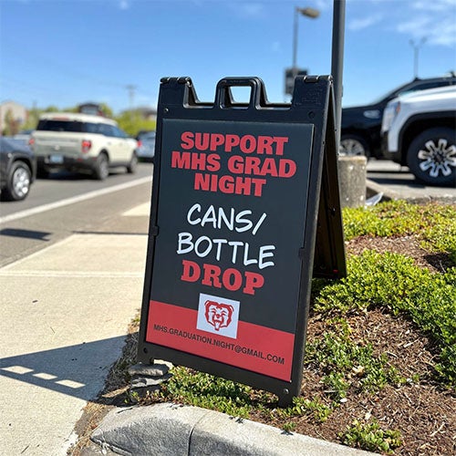 A-frame sign advertising 'Support MHS Grad Night Cans/Bottle Drop' and an email address next to a street.