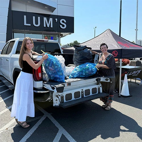 Two women stand by a truck bed filled with bags of cans, recycled outside LUM'S' building.