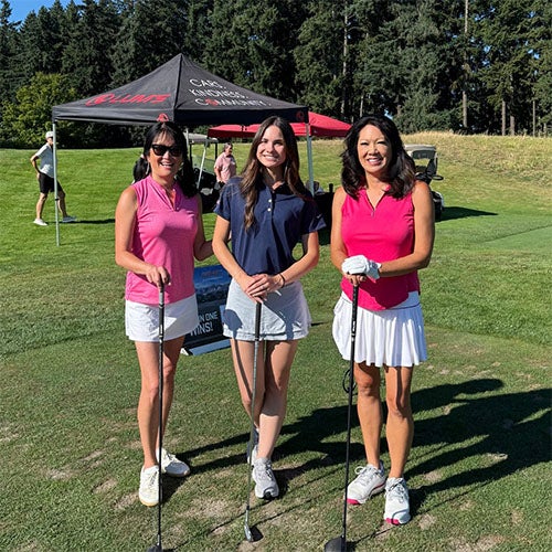 Three women in golf attire holding clubs pose on a golf course with tents and trees in the background.