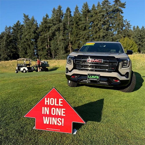 A red arrow sign that says 'HOLE IN ONE WINS!' points to a light-colored GMC truck on a golf course.