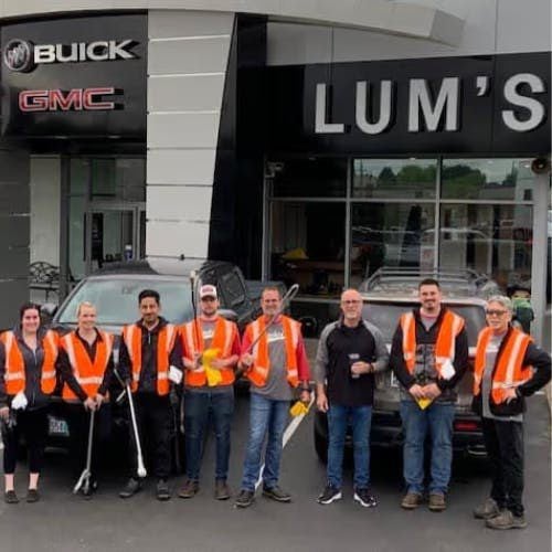 Group of 8 people, some in orange safety vests, stand in front of a car dealership, possibly after a cleanup.