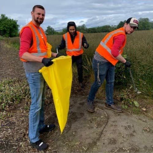 Three people wearing orange safety vests and gloves pick up litter along a dirt path in a grassy field.