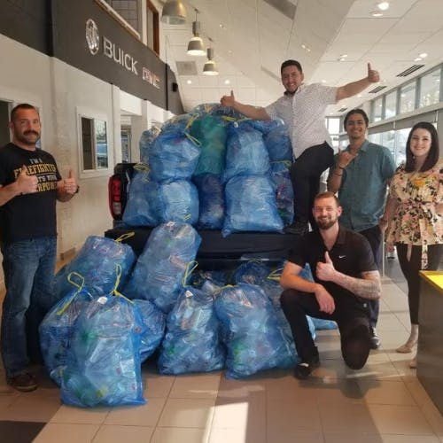 A group of five people, four men and one woman, stand around a truck bed filled with bags of plastic bottles.