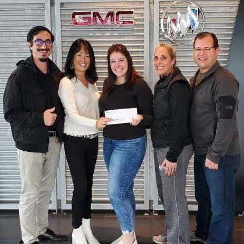 Five people, three women and two men, standing and smiling in front of GMC and Buick logos. The woman in the middle holds a white envelope.