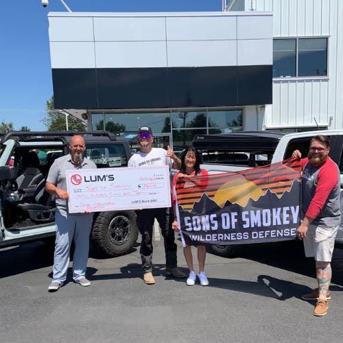 Four people hold a large check and 'Sons of Smokey' banner while standing next to vehicles.