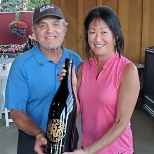A man and woman hold a large wine bottle. The man wears a blue polo and a 'US Open' cap. The woman wears a pink top.