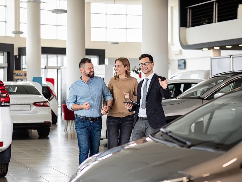 A smiling couple walks through a car dealership, holding hands, with a salesman showing them cars.